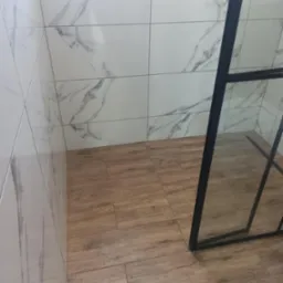 Wykończona kabina prysznicowa z drewnopodobną podłogą i białymi płytkami imitującymi marmur na ścianach. Czarna rama kabiny prysznicowej.