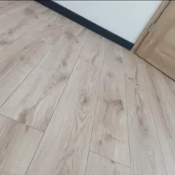 Jasna podłoga panelowa ułożona pod kątem, z widoczną przypodłogową listwą w kolorze czarnym i fragmentem drewnianych drzwi z czarnym akcentem.