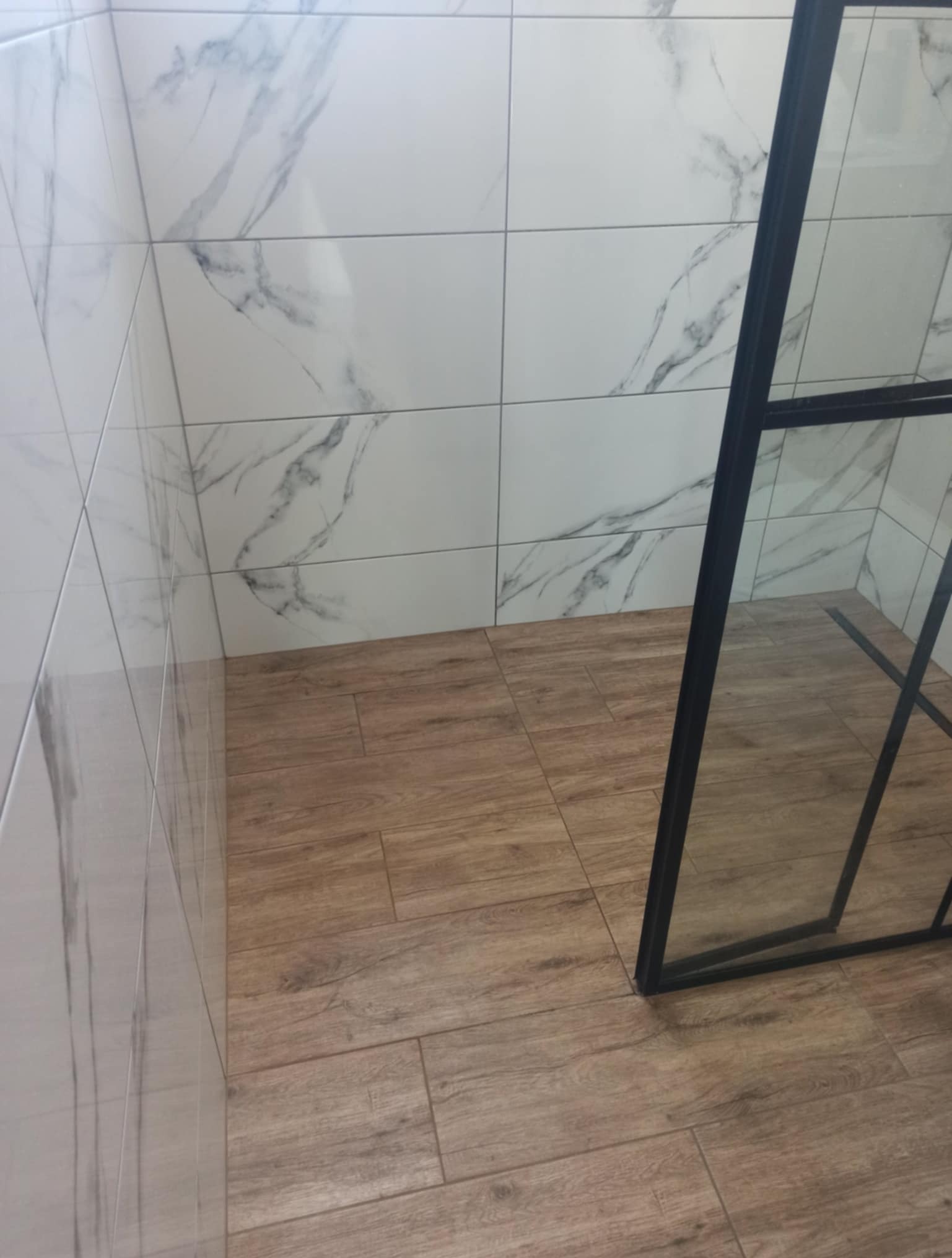 Wykończona kabina prysznicowa z drewnopodobną podłogą i białymi płytkami imitującymi marmur na ścianach. Czarna rama kabiny prysznicowej.