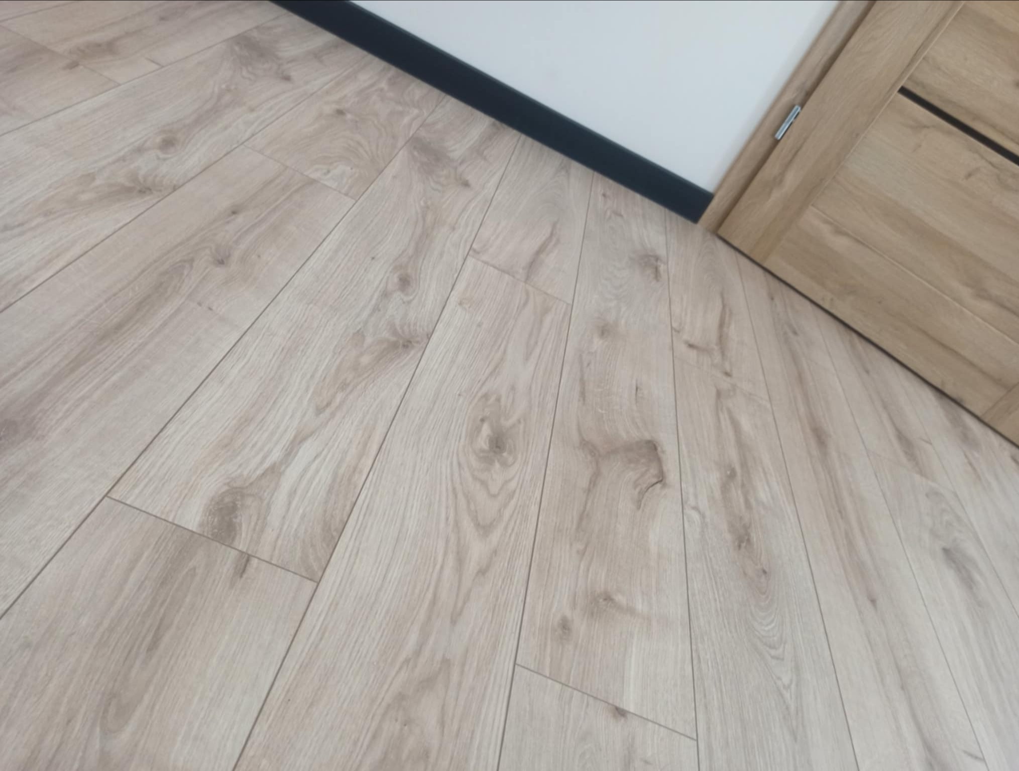 Jasna podłoga panelowa ułożona pod kątem, z widoczną przypodłogową listwą w kolorze czarnym i fragmentem drewnianych drzwi z czarnym akcentem.