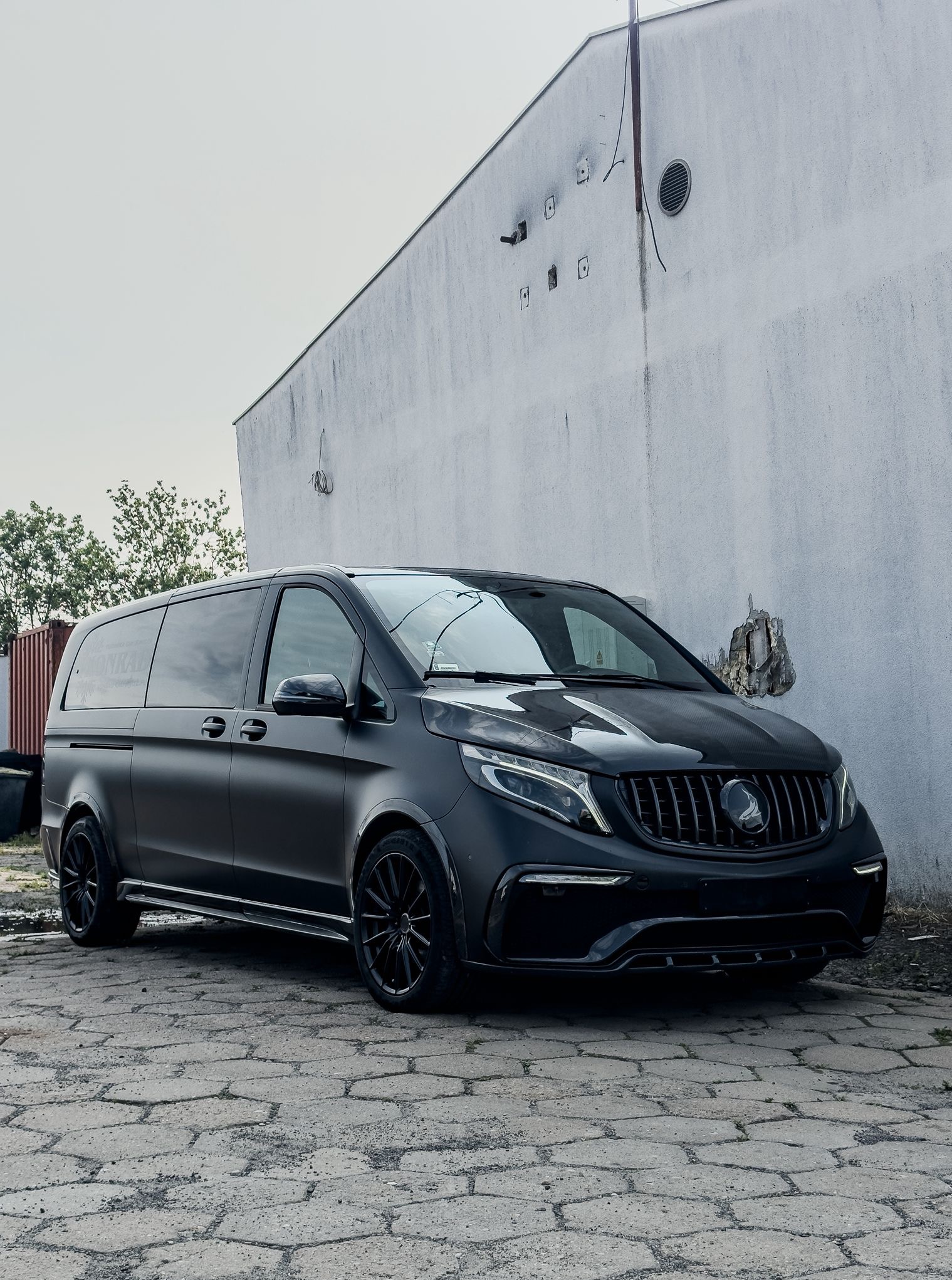 Czarny, matowy van Mercedes z niestandardowym grillem stoi na tle białej ściany. Widoczne detale tuningu nadwozia i felgi. Auto po oklejeniu folią.