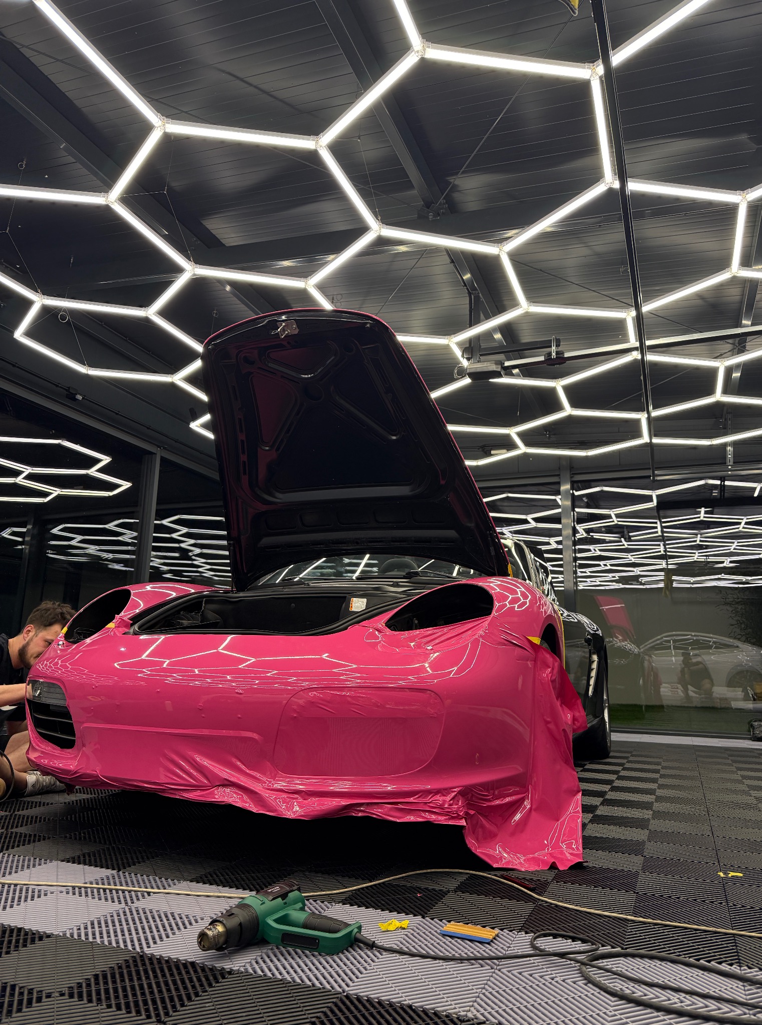 Różowy sportowy samochód w trakcie oklejania folią. Otwarta maska, widoczne narzędzia i pracownik. Charakterystyczne oświetlenie sufitowe.