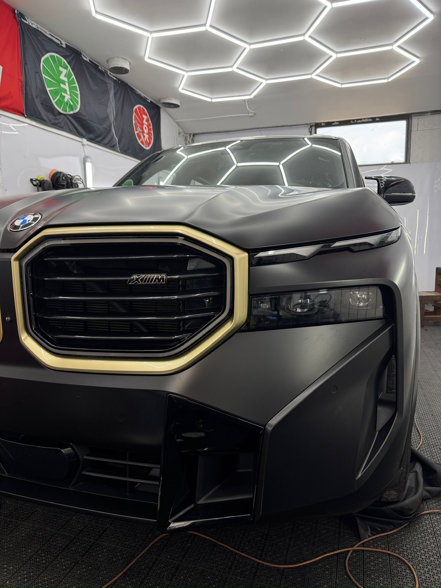 Matowy czarny BMW X7 z złotymi akcentami na grillu, widziany z bliska w dobrze oświetlonym warsztacie. Detale karoserii i oświetlenia sufitowego w kształcie plastra miodu.
