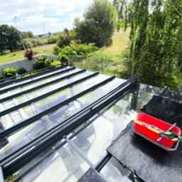 Mycie szklanego dachu oranżerii z użyciem czerwonego wiadra z narzędziami. Widok z góry na czyszczoną powierzchnię i otaczającą zieleń. Przejrzystość szkła po umyciu.