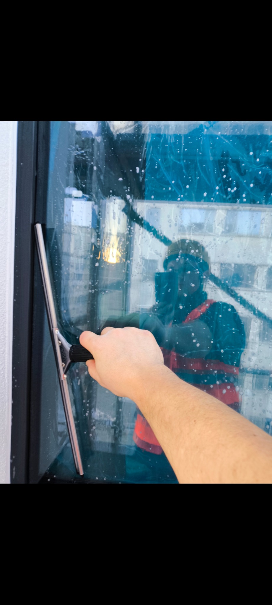 Czyszczenie okna z zewnątrz za pomocą ściągaczki, widoczne krople wody i odbicie osoby w kamizelce odblaskowej.