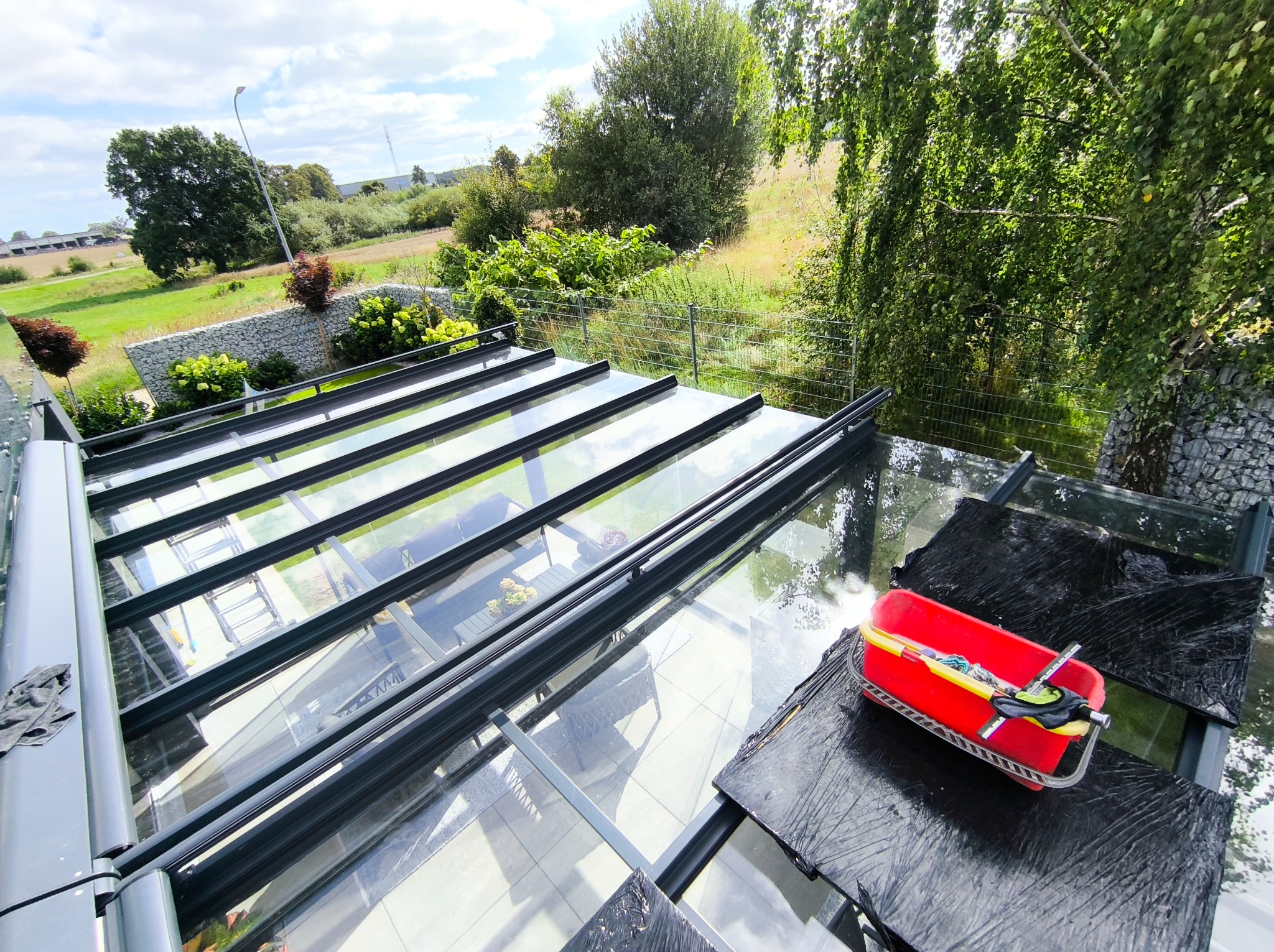 Mycie szklanego dachu oranżerii z użyciem czerwonego wiadra z narzędziami. Widok z góry na czyszczoną powierzchnię i otaczającą zieleń. Przejrzystość szkła po umyciu.