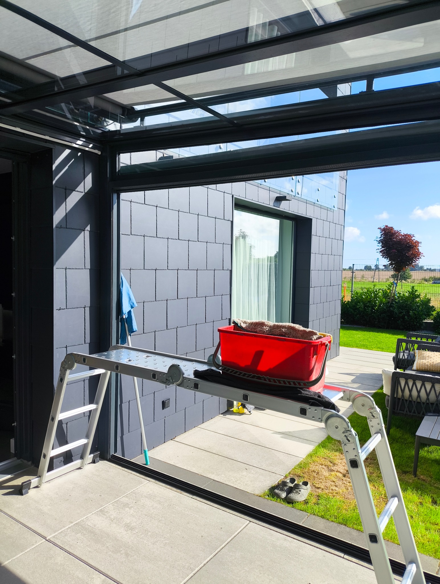 Rozstawiona drabina teleskopowa z czerwonym wiadrem i mopem na tarasie domu z szarą elewacją i oknami. Widok na zielony ogród.