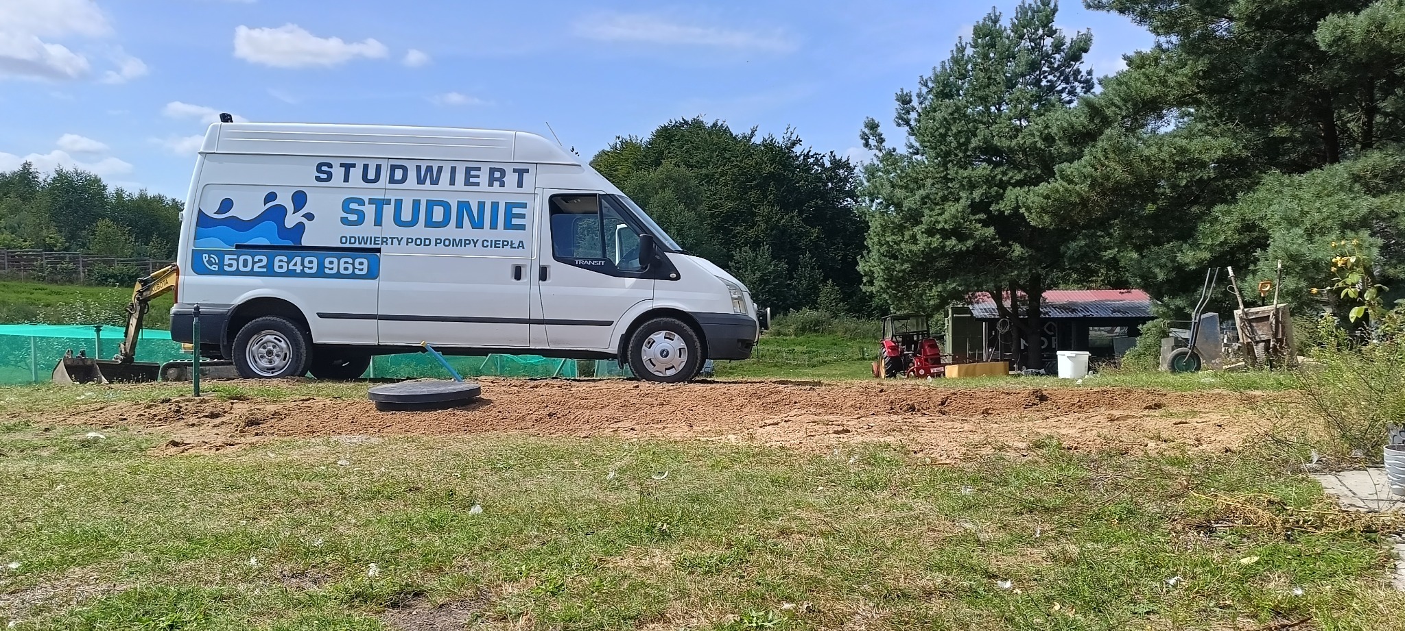 Biały bus z logo firmy wiertniczej na tle wykopu i zieleni. Widoczny mały traktor i elementy studni. Dzień słoneczny, błękitne niebo.