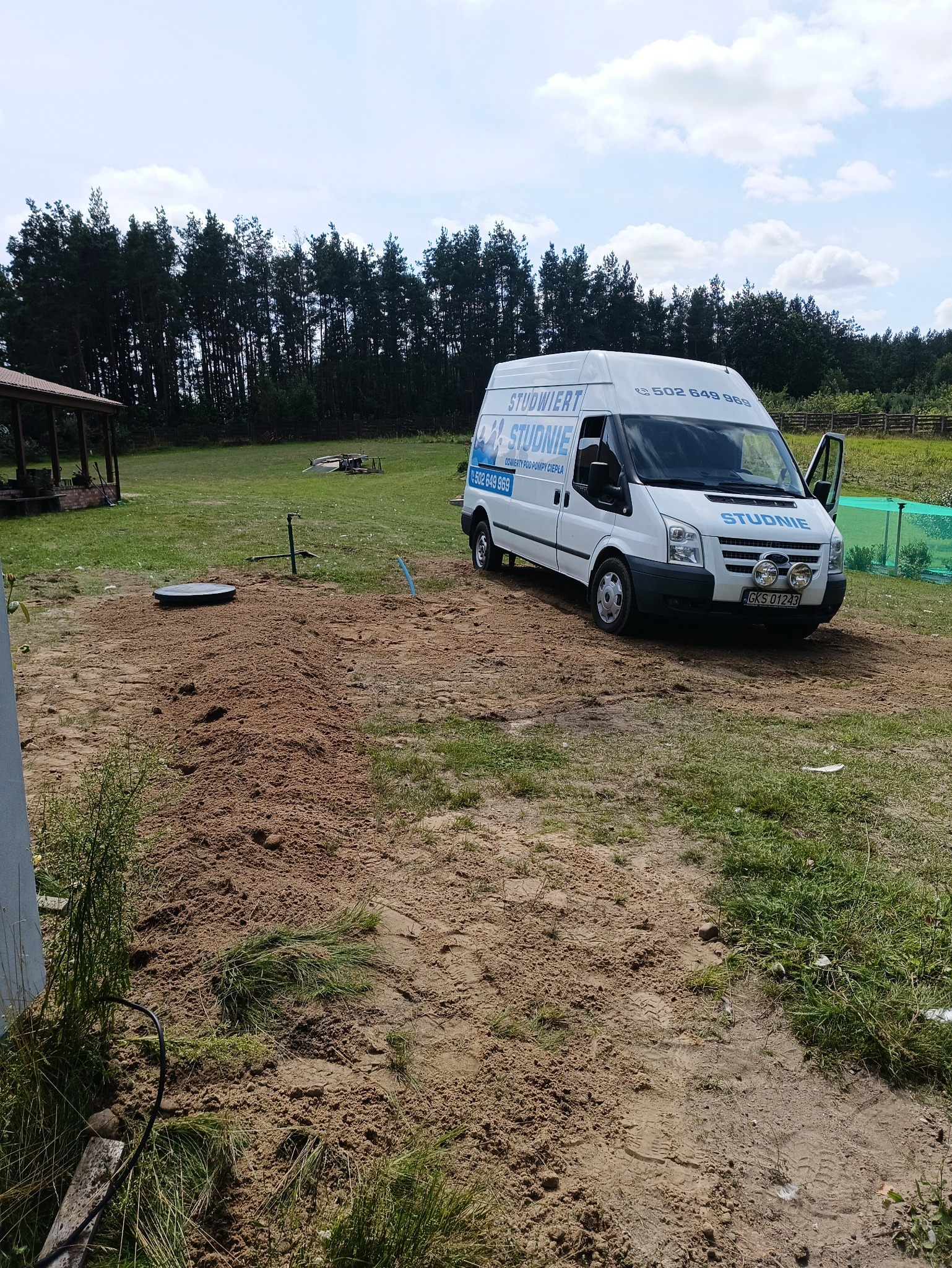 Wykonany wykop pod studnię kręgową na działce, obok zaparkowany biały bus firmy z otwartymi drzwiami. W tle las i zabudowania gospodarcze.