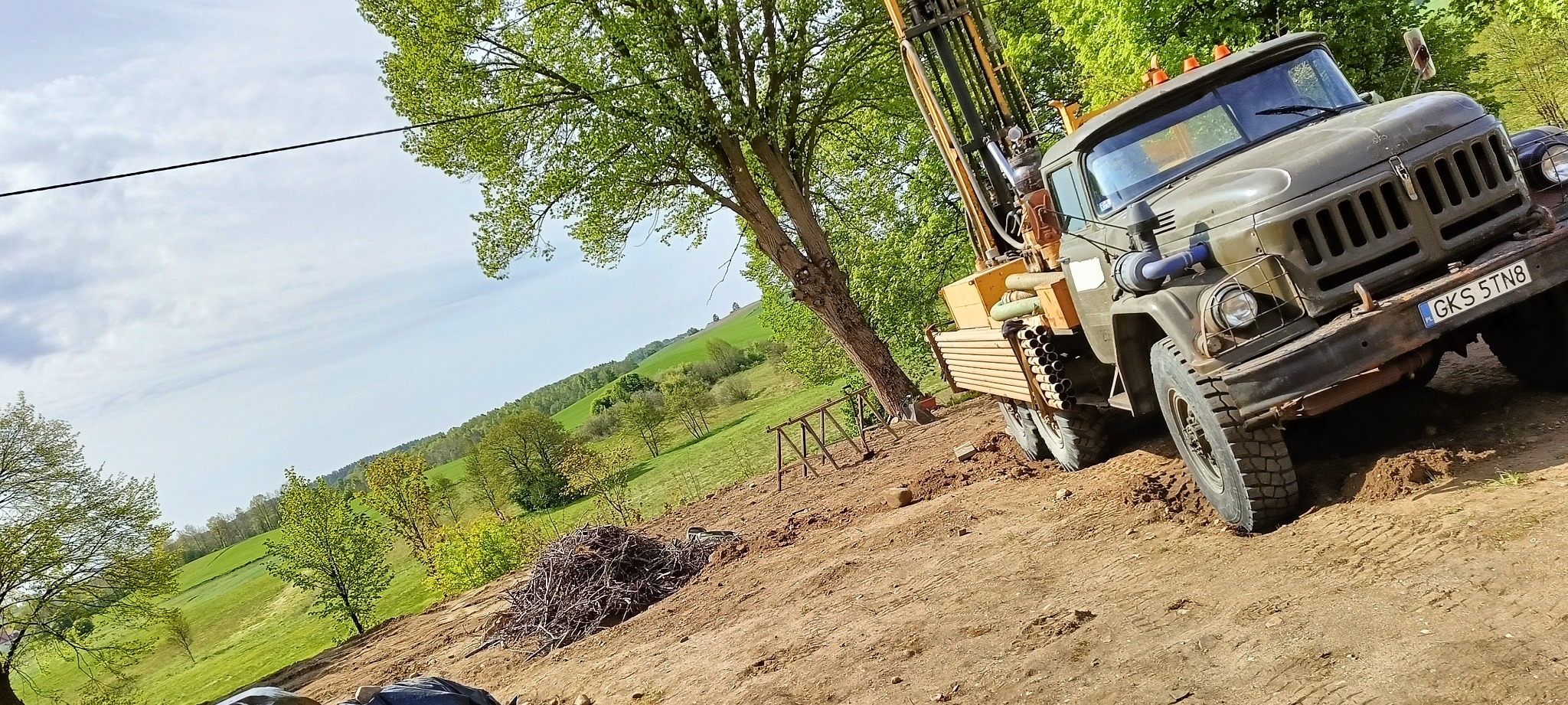 Żuraw wiertniczy na nierównym terenie, z widocznymi rurami wiertniczymi i zielonym krajobrazem w tle. Ujęcie z niskiego kąta, podkreślające potęgę maszyny.
