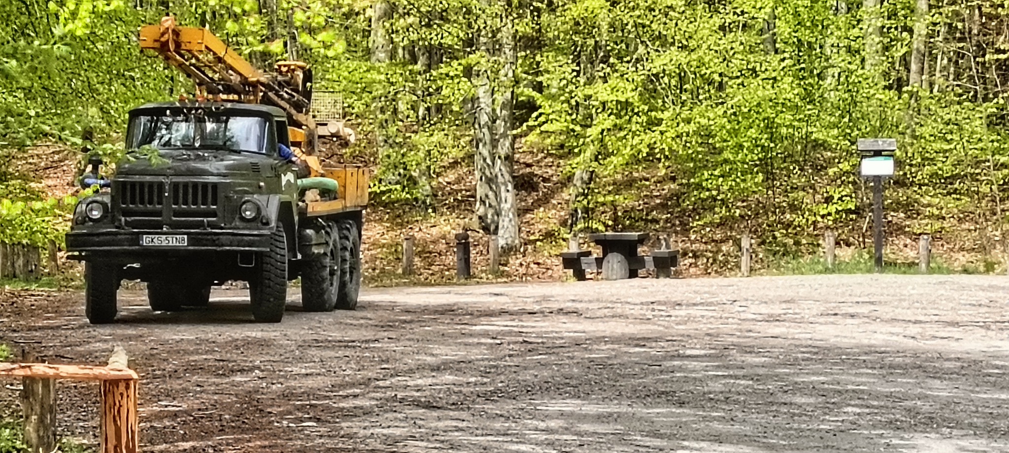 Ciężarówka z wiertnicą na tle leśnym. Wiertnica zamontowana na pojeździe sugeruje wiercenie studni. Widoczny fragment tablicy informacyjnej.