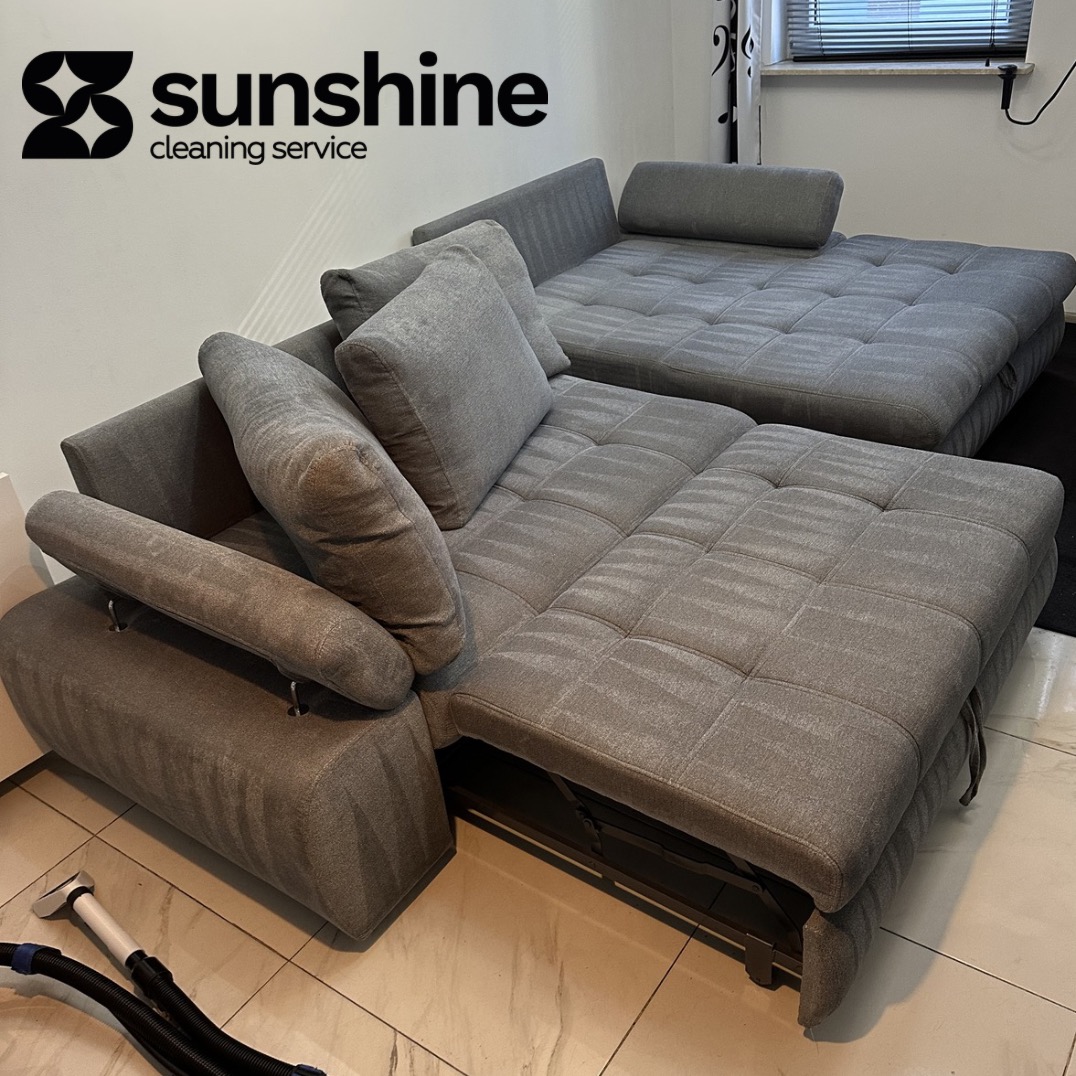Szara, rozłożona sofa modułowa z poduszkami po czyszczeniu, widoczny fragment odkurzacza piorącego. Logo firmy w rogu kadru.