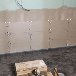 Szakul Bud - Układanie płytek na ścianie z użyciem krzyżyków dystansowych i poziomicy laserowej. Widoczna paczka płytek i folia ochronna na podłodze.
