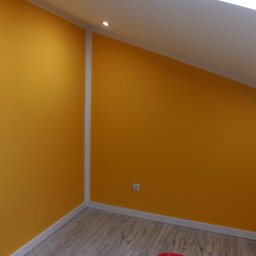 Szakul Bud - Żółta ściana w pokoju na poddaszu z oknem dachowym i drewnianą podłogą. Listwy przypodłogowe i narożnikowe w kolorze białym. Widoczny fragment gniazdka.