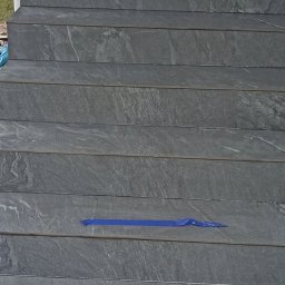 Szakul Bud - Wykończone schody zewnętrzne z szarych płytek imitujących kamień, zabezpieczone niebieską taśmą malarską na kilku stopniach. Widok z dołu, z fragmentem osoby w tle.