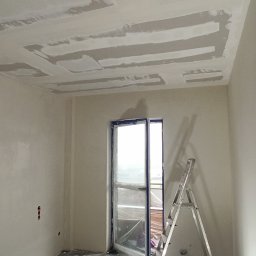 Szakul Bud - Pokój w trakcie prac wykończeniowych: szpachlowane ściany i sufit z widocznymi łączeniami płyt gipsowo-kartonowych, drabina oparta o okno balkonowe.
