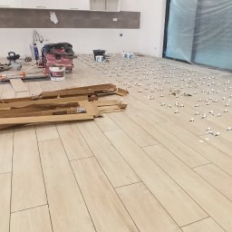 Szakul Bud - Układanie płytek podłogowych w dużym pomieszczeniu. Widoczne narzędzia glazurnicze, płytki, dystanse i puste kartony po płytkach. Jasne, naturalne światło.
