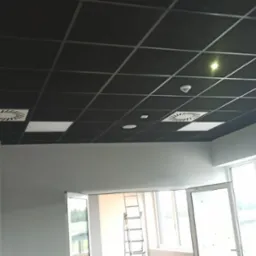 Kwadratowy, czarny sufit podwieszany z kratownicą, oświetleniem i wentylacją. Klimatyzatory w folii ochronnej na ścianie, białe ściany i okna, drabina w tle.