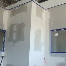 Szakul Bud - Wnętrze w trakcie szpachlowania: ściany z nałożoną gładzią, zabezpieczone okna i sufit taśmą malarską, widoczny klimatyzator i drabina malarska.
