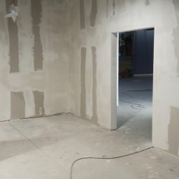Szakul Bud - Wnętrze w trakcie szpachlowania: ściany z płyt gipsowo-kartonowych z widocznymi paskami szpachli, surowa podłoga, widoczne kable i instalacje pod sufitem.