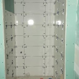 Szakul Bud - Montaż białych płytek wnęki z użyciem klipsów poziomujących. Ściany wykończone płytami karton-gips, na podłodze widoczne narzędzia i wiadro z zaprawą.