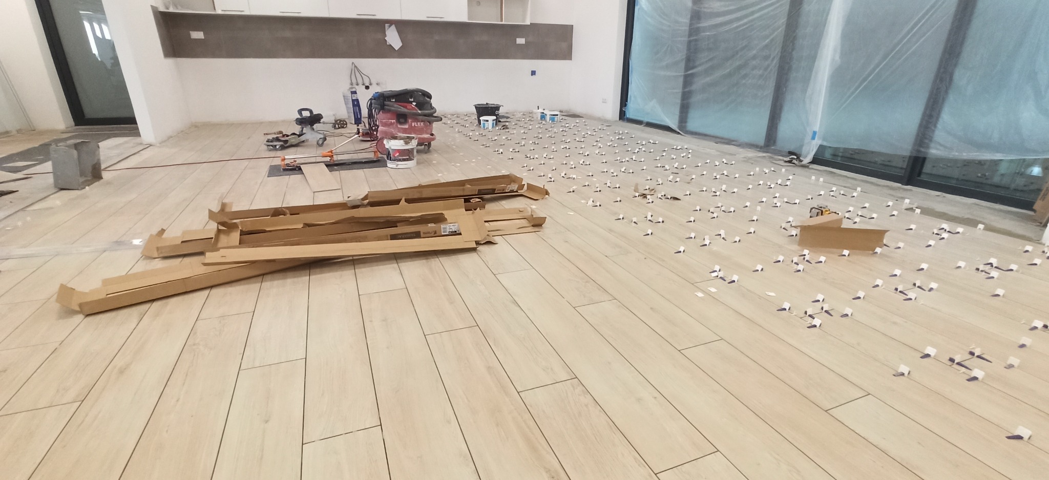 Układanie jasnych płytek podłogowych z użyciem krzyżyków dystansowych. Widoczne narzędzia glazurnicze i opakowania po płytkach. Duże okno w tle.