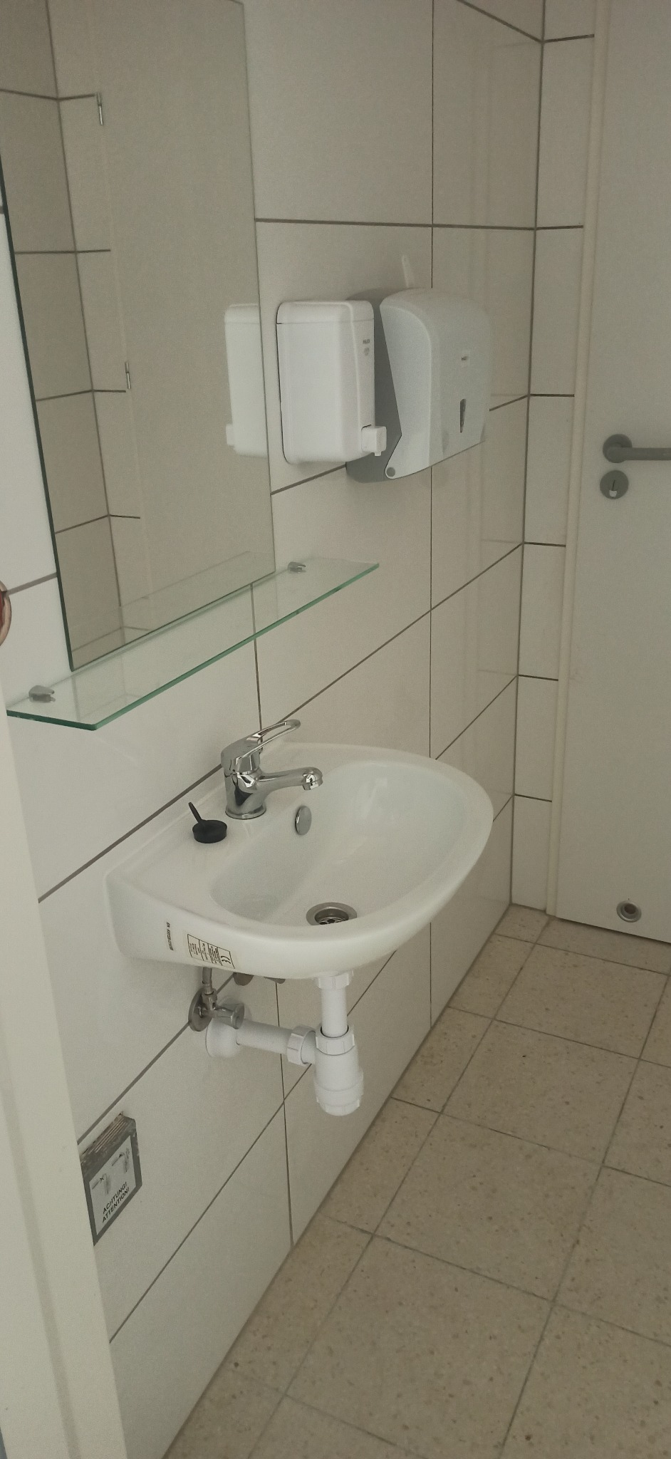 Biała umywalka zamontowana na ścianie z jasnymi płytkami, z baterią i szklaną półką pod lustrem. Widoczne dozowniki mydła i ręczników oraz fragment drzwi.