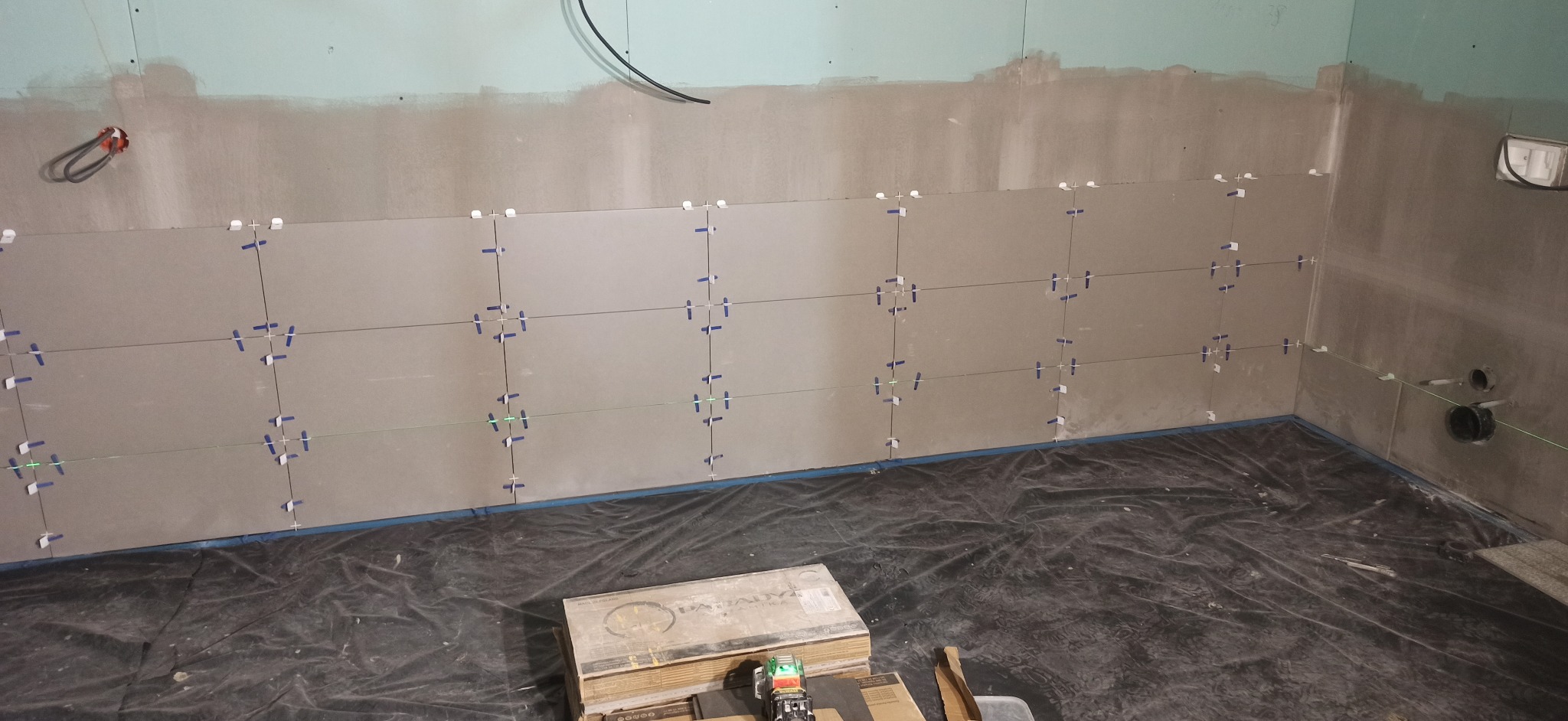 Układanie płytek na ścianie z użyciem krzyżyków dystansowych i poziomicy laserowej. Widoczna paczka płytek i folia ochronna na podłodze.