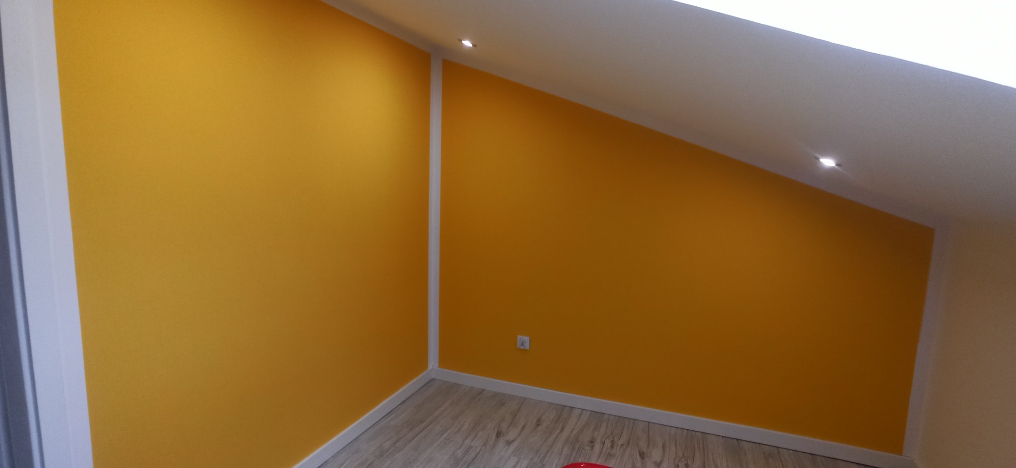 Żółta ściana w pokoju na poddaszu z oknem dachowym i drewnianą podłogą. Listwy przypodłogowe i narożnikowe w kolorze białym. Widoczny fragment gniazdka.