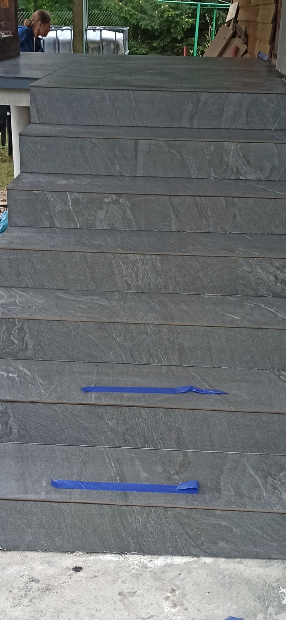 Wykończone schody zewnętrzne z szarych płytek imitujących kamień, zabezpieczone niebieską taśmą malarską na kilku stopniach. Widok z dołu, z fragmentem osoby w tle.