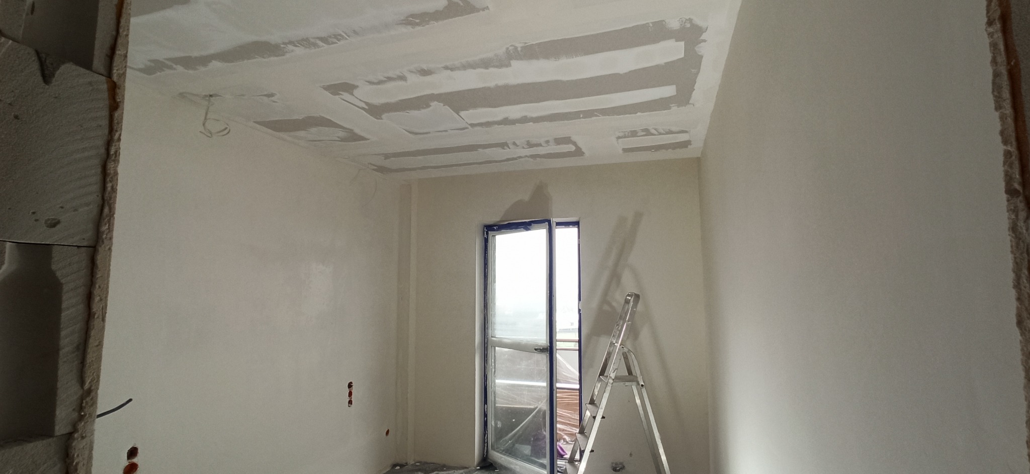 Pokój w trakcie prac wykończeniowych: szpachlowane ściany i sufit z widocznymi łączeniami płyt gipsowo-kartonowych, drabina oparta o okno balkonowe.