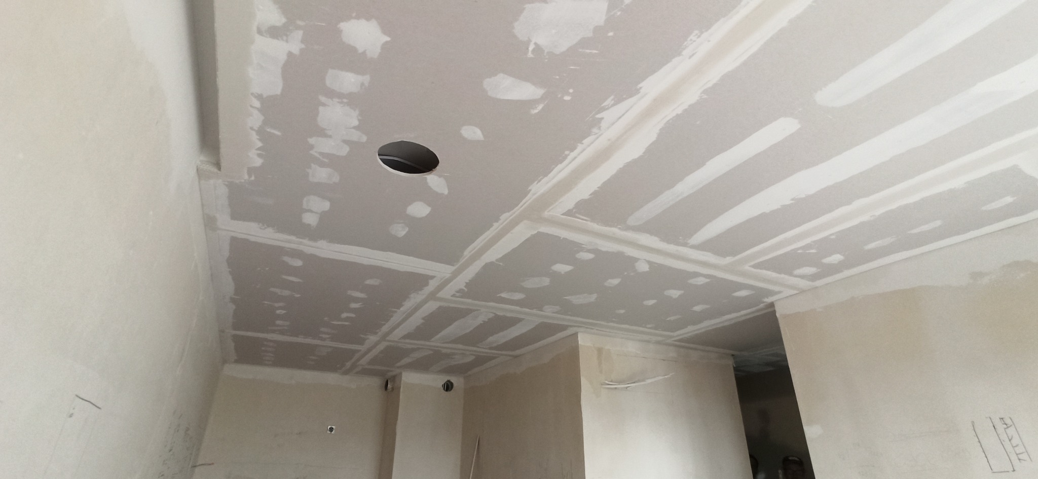 Sufit z płyt gipsowo-kartonowych w trakcie szpachlowania, widoczne łączenia i otwór na oświetlenie. Ściany przygotowane do dalszych prac wykończeniowych.