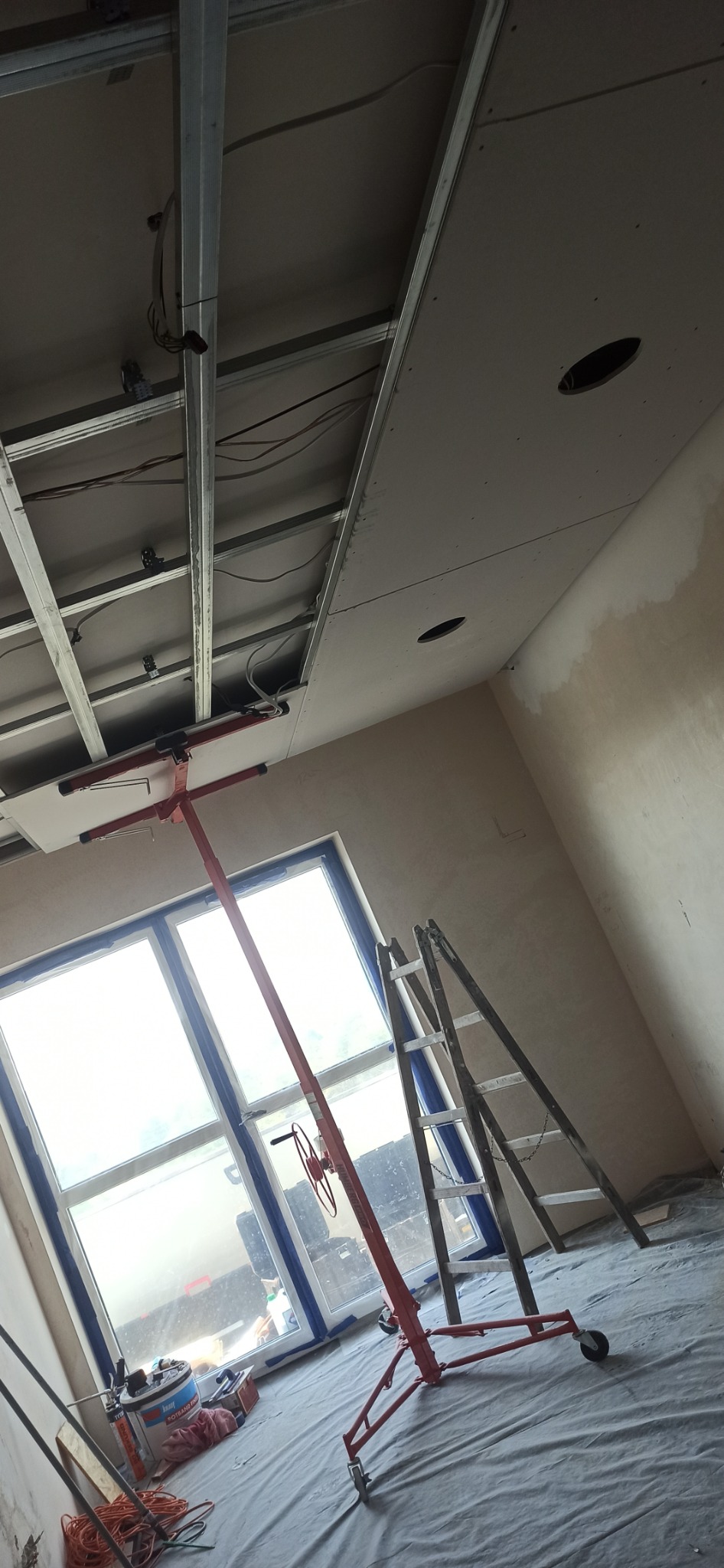 Montaż sufitu podwieszanego z płyty g-k, widoczny stelaż i podnośnik. Okno z niebieską folią zabezpieczającą, drabina. Prace wykończeniowe w nowym budynku.