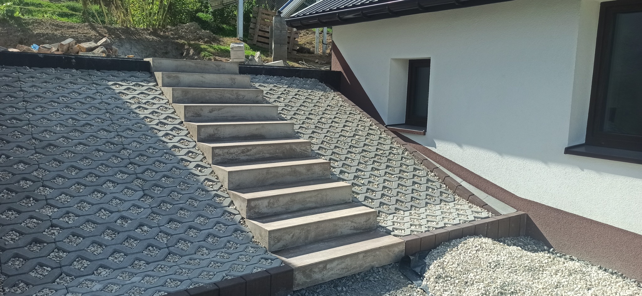 Schody zewnętrzne z szarej kostki ażurowej i betonowych stopni prowadzące wzdłuż białej ściany budynku z oknem, wykończone drobnym kamieniem.