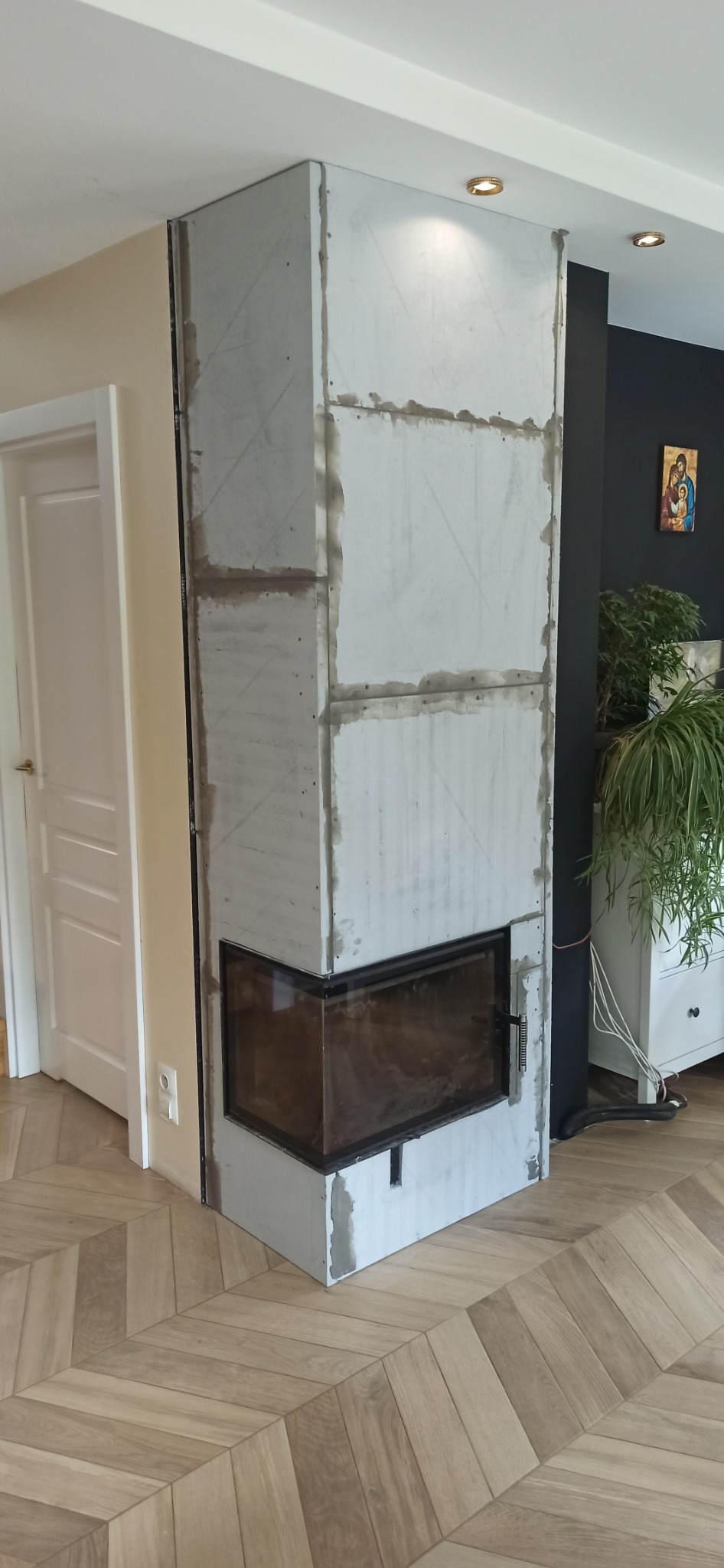 Wnętrze z kominkiem obudowanym płytami, widoczne ślady szpachlowania. Parkiet ułożony w jodełkę, białe drzwi w tle. Minimalistyczny wystrój.