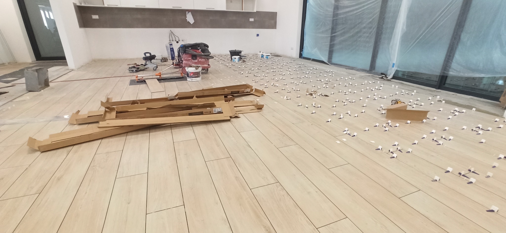 Układanie płytek podłogowych w dużym pomieszczeniu. Widoczne narzędzia glazurnicze, płytki, dystanse i puste kartony po płytkach. Jasne, naturalne światło.