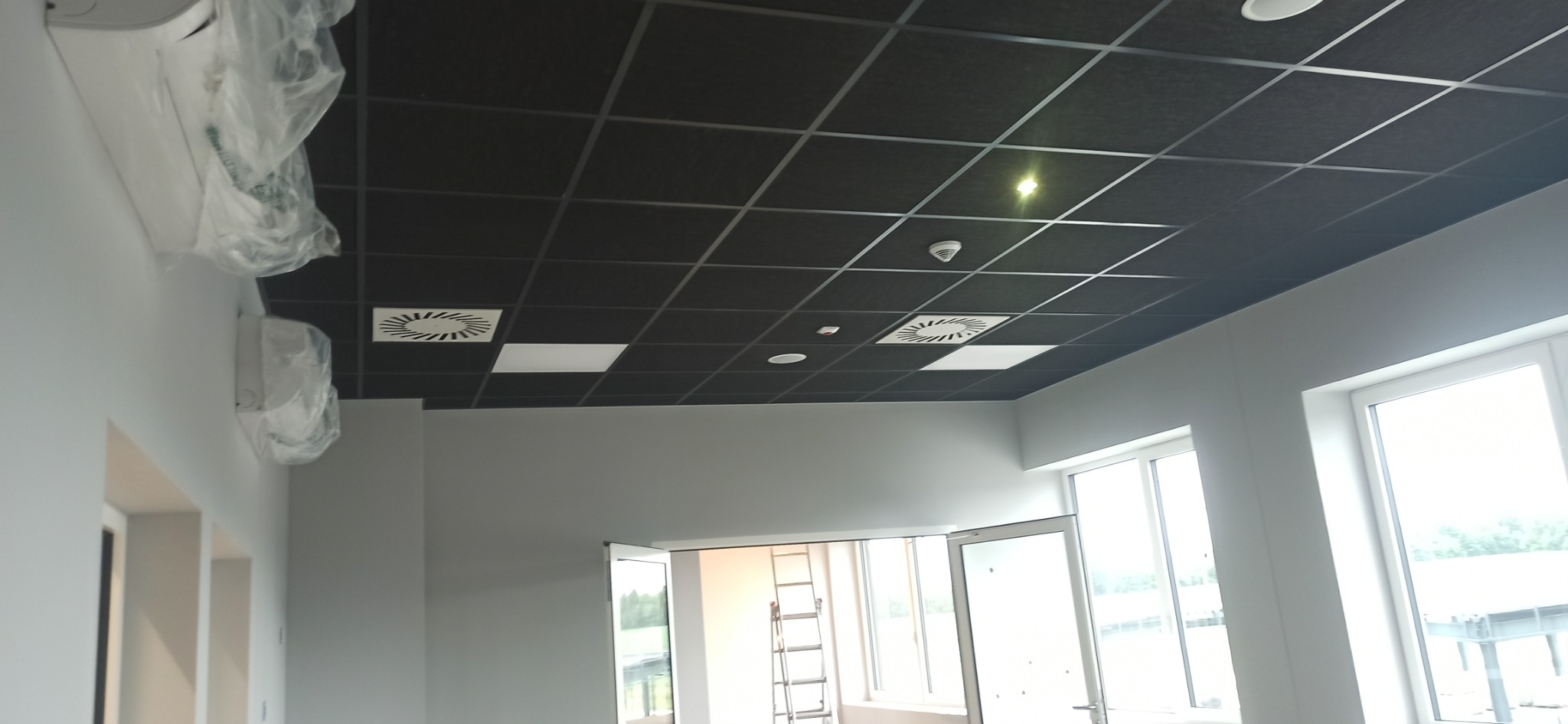 Kwadratowy, czarny sufit podwieszany z kratownicą, oświetleniem i wentylacją. Klimatyzatory w folii ochronnej na ścianie, białe ściany i okna, drabina w tle.