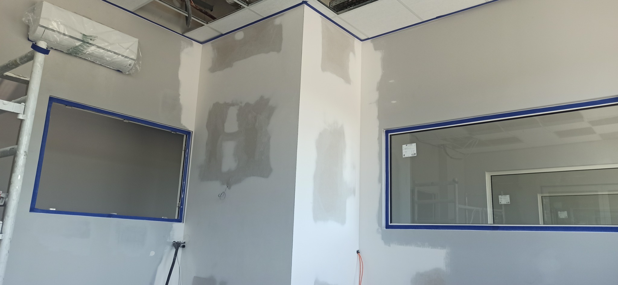 Wnętrze w trakcie szpachlowania: ściany z nałożoną gładzią, zabezpieczone okna i sufit taśmą malarską, widoczny klimatyzator i drabina malarska.