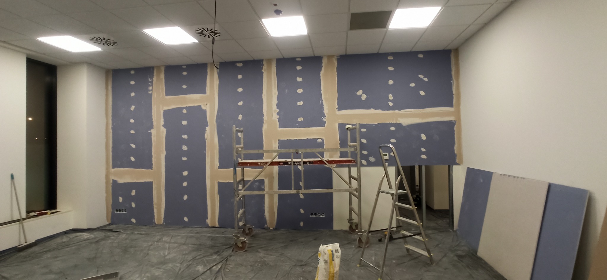 Wnętrze w trakcie prac z płytami gipsowo-kartonowymi, widoczne rusztowanie i drabina. Ściany częściowo pokryte szpachlą, sufit z oświetleniem rastrowym.