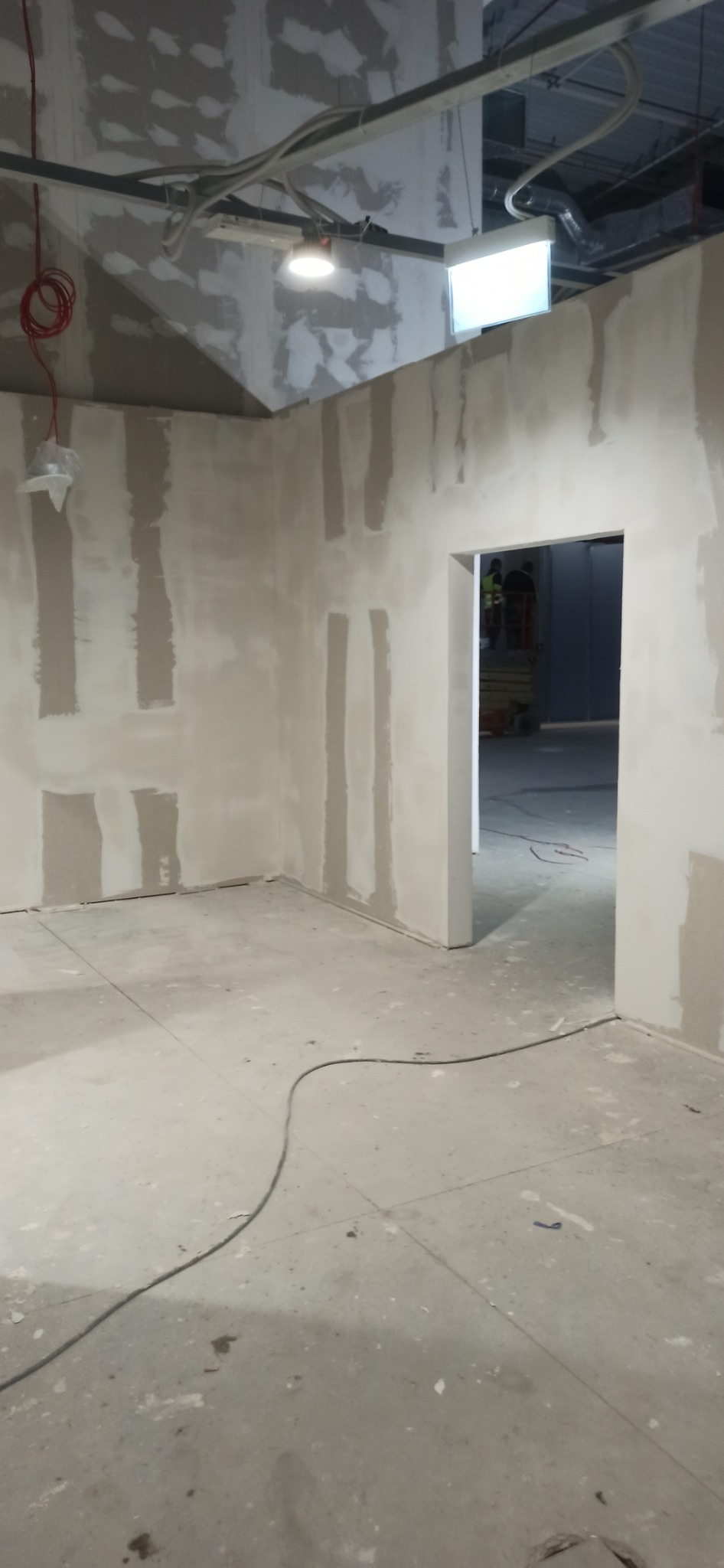 Wnętrze w trakcie szpachlowania: ściany z płyt gipsowo-kartonowych z widocznymi paskami szpachli, surowa podłoga, widoczne kable i instalacje pod sufitem.