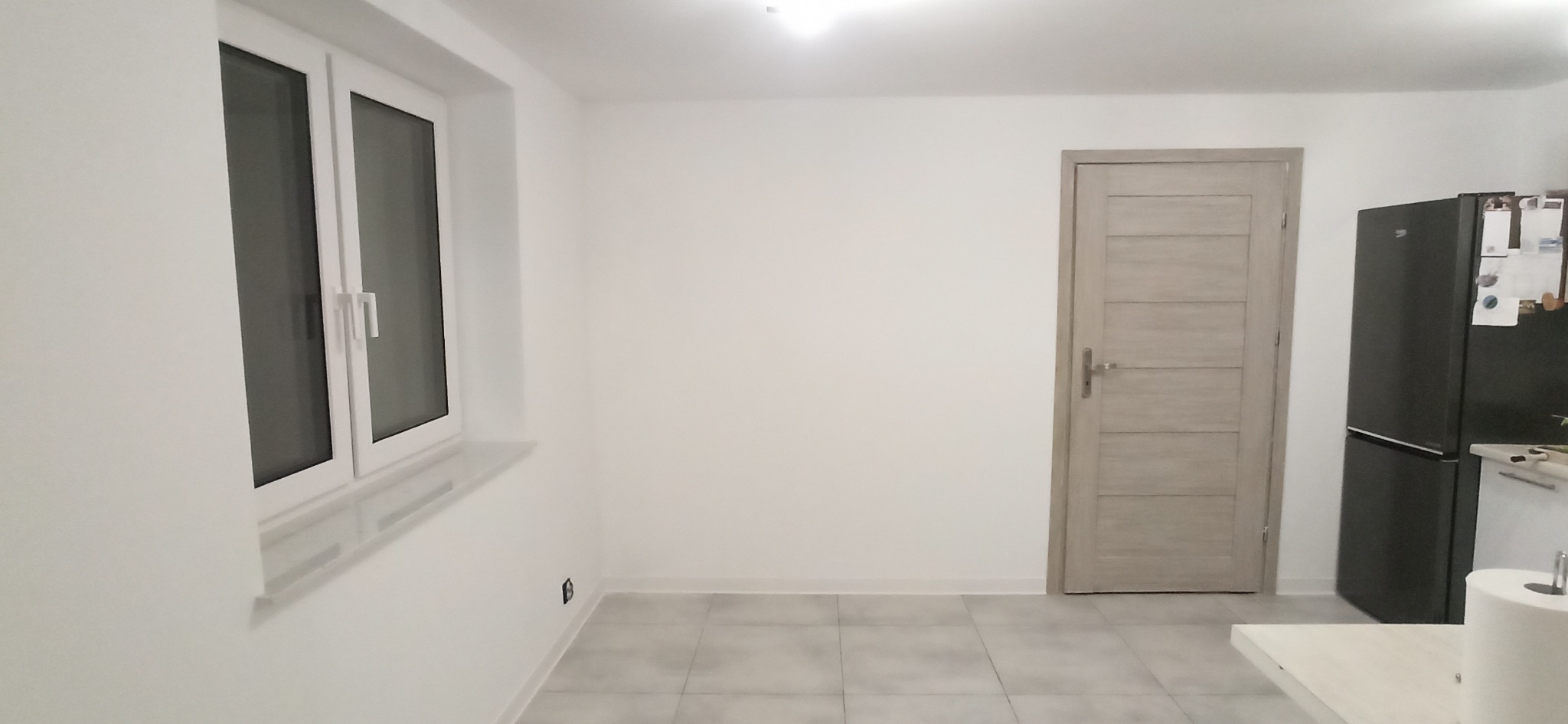 Jasne wnętrze po remoncie: białe ściany, okno PCV, szara podłoga, drewniane drzwi i czarna lodówka. Minimalistyczny design.