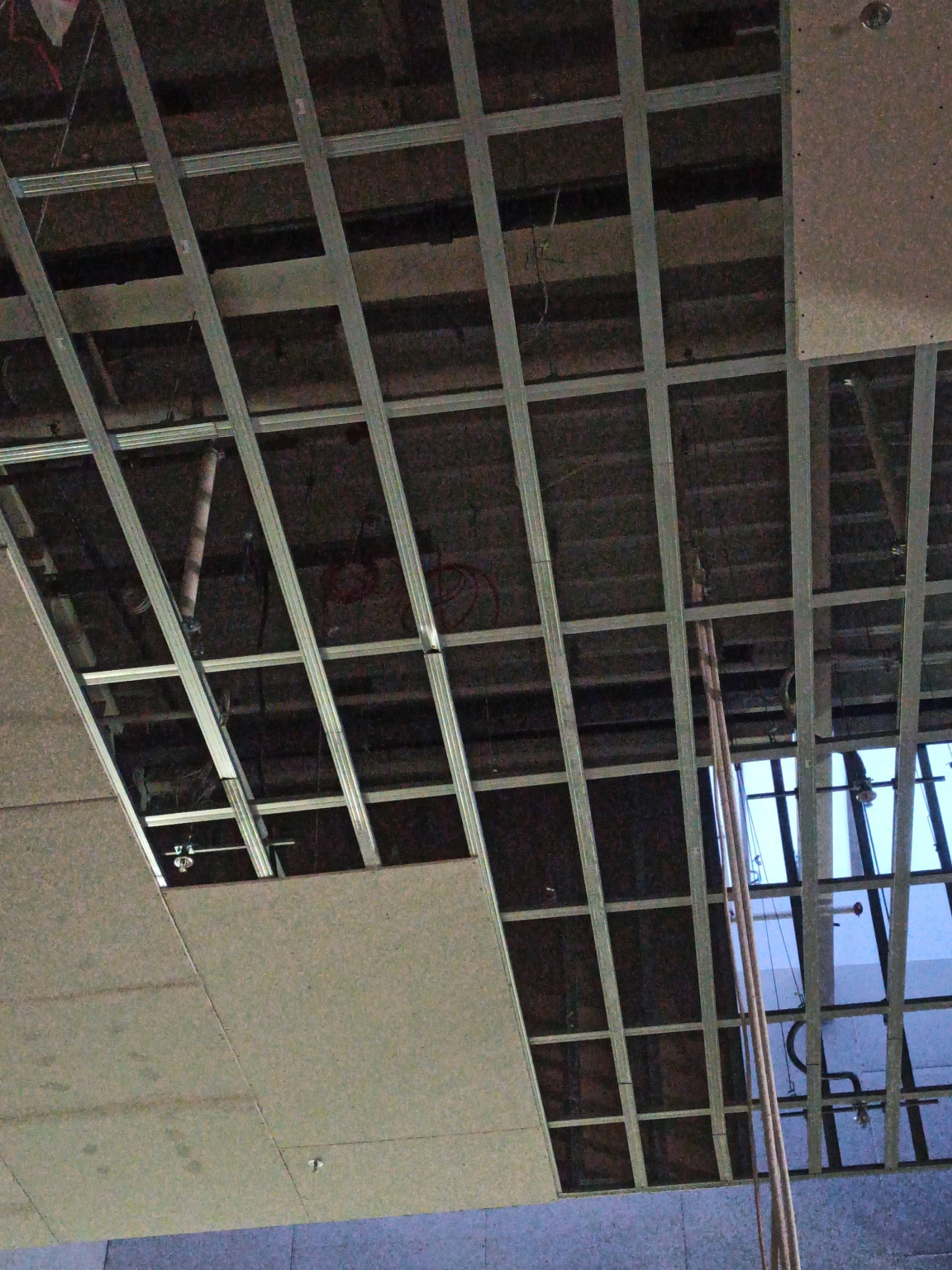 Metalowa konstrukcja sufitu podwieszanego z częściowo zamontowanymi płytami. Widoczne instalacje i okno w tle. Ujęcie z dołu.