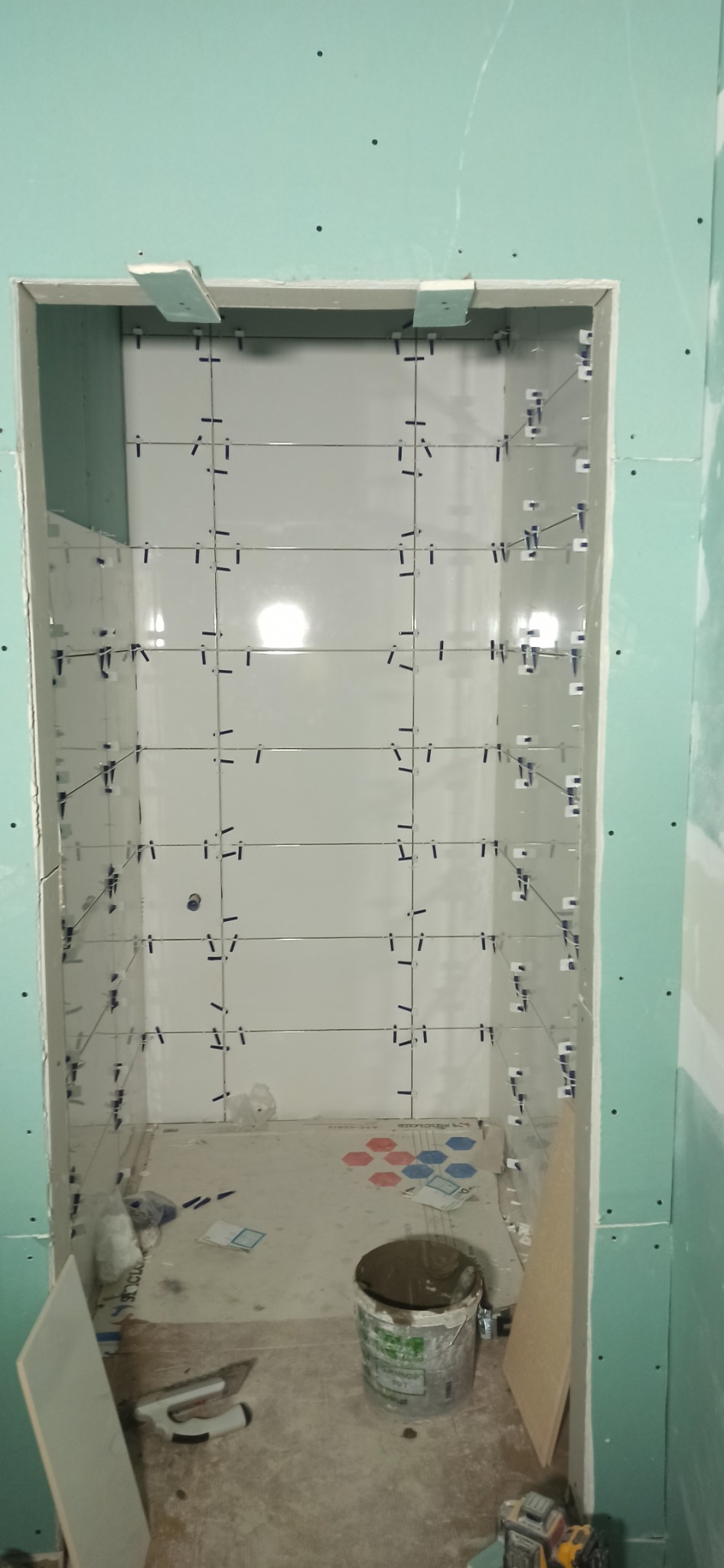 Montaż białych płytek wnęki z użyciem klipsów poziomujących. Ściany wykończone płytami karton-gips, na podłodze widoczne narzędzia i wiadro z zaprawą.