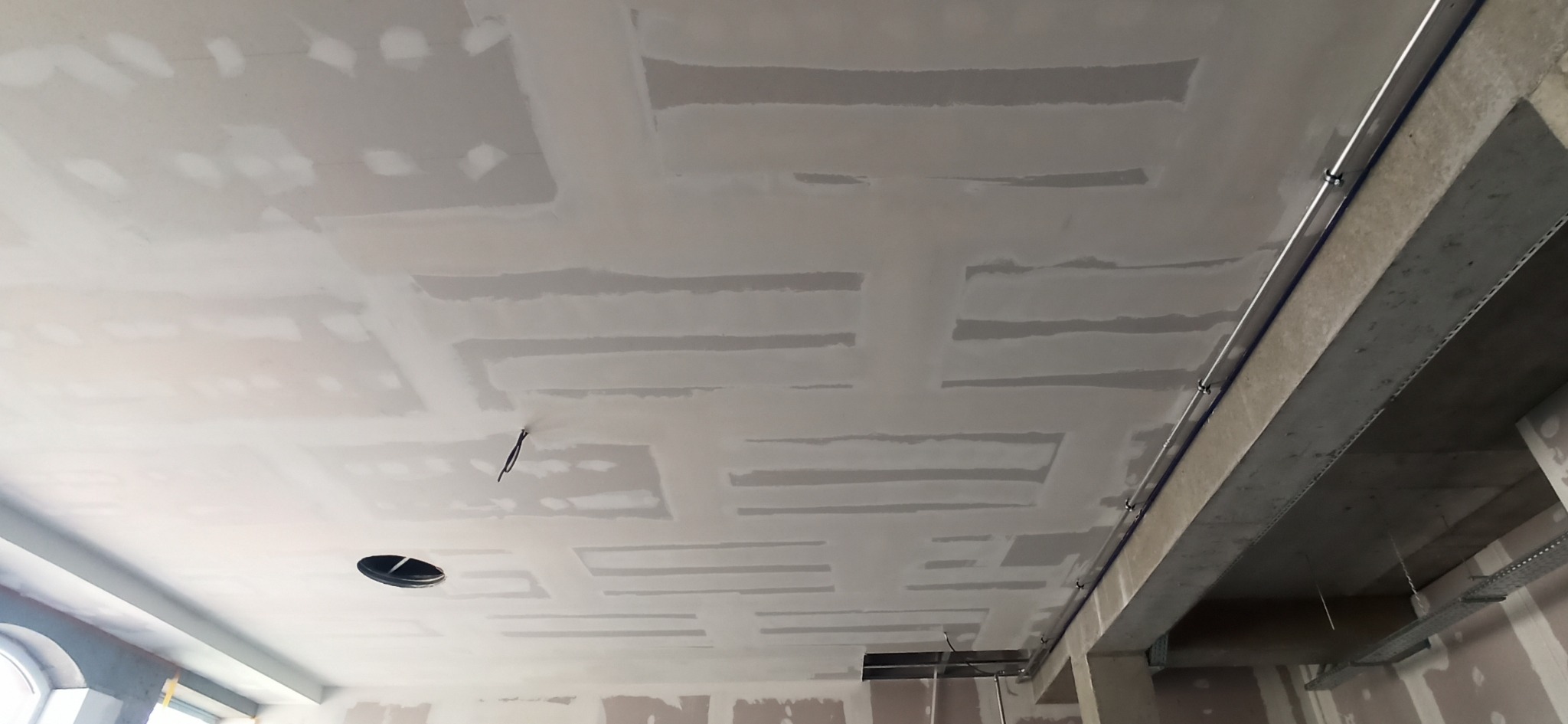 Sufit z płyt kartonowo-gipsowych, częściowo wyszpachlowany, widoczne łączenia i otwór na oświetlenie. Szara kolorystyka, surowy betonowy element konstrukcyjny.