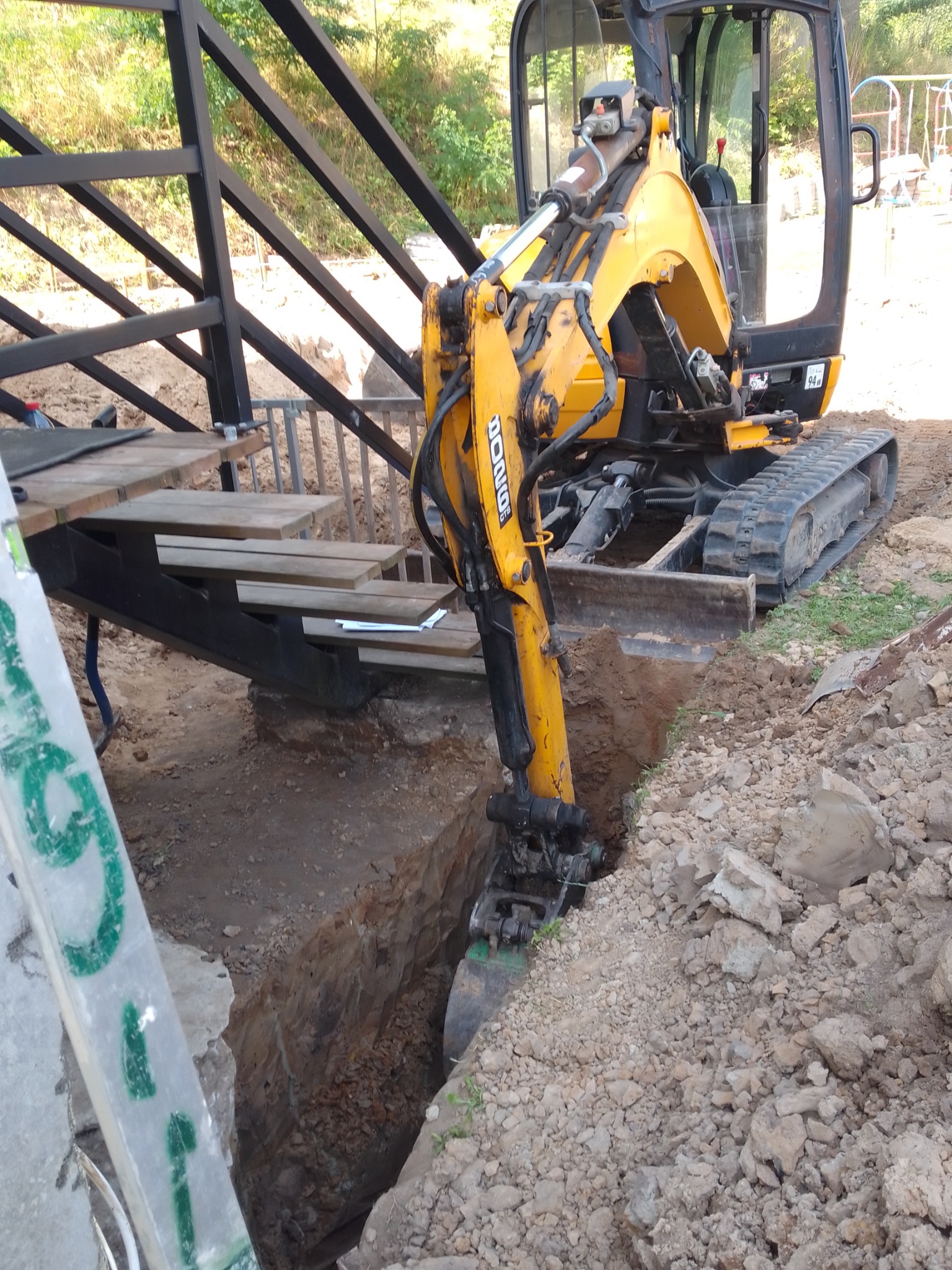 Żółta minikoparka JCB w trakcie wykopu wąskiego rowu obok stalowej konstrukcji schodów. Widoczne ślady pracy i usypana ziemia.