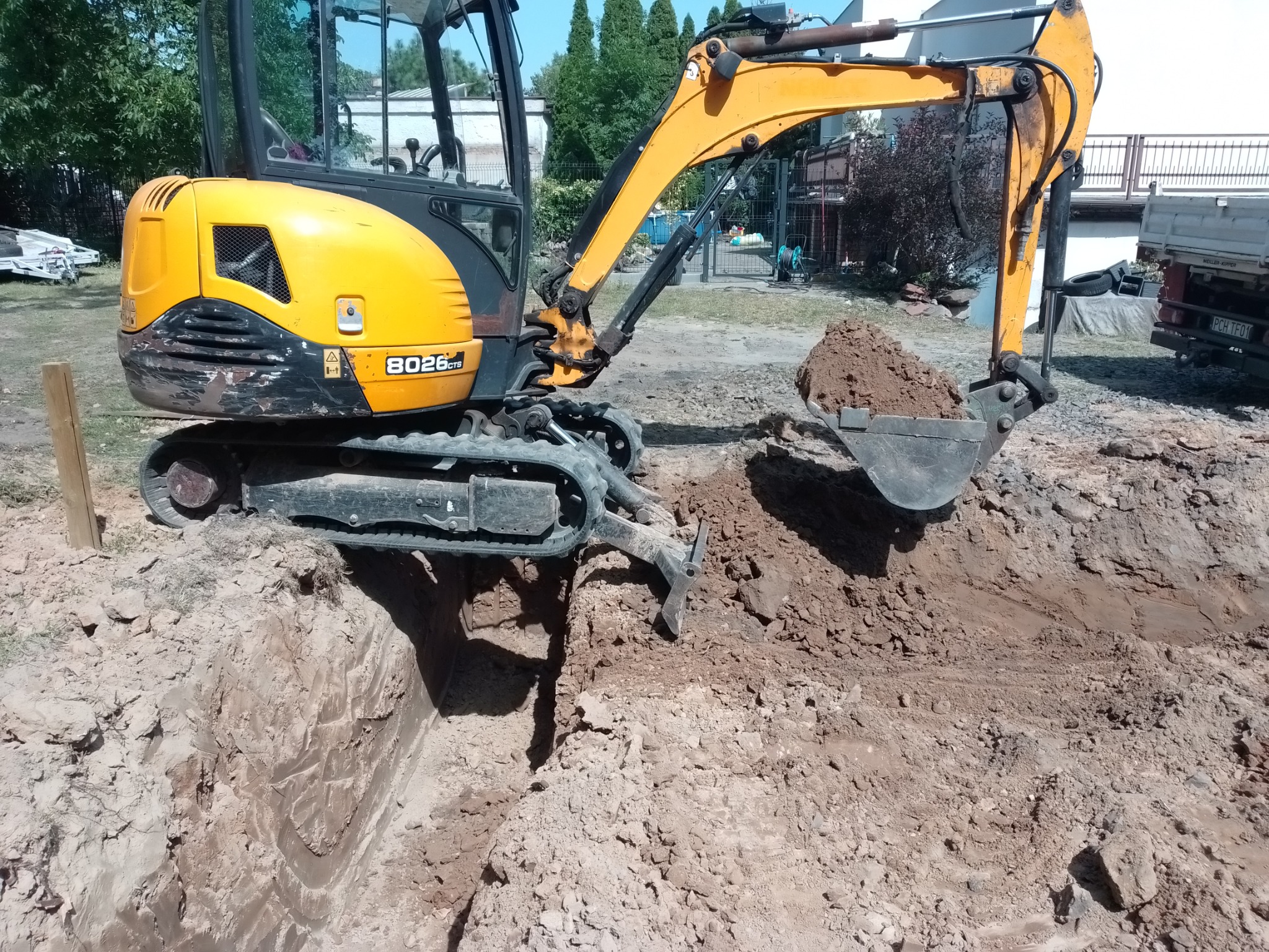 Żółta minikoparka JCB 8026 CTS z pełną łyżką ziemi na wykopie, widok z boku, częściowo widoczny samochód ciężarowy w tle.