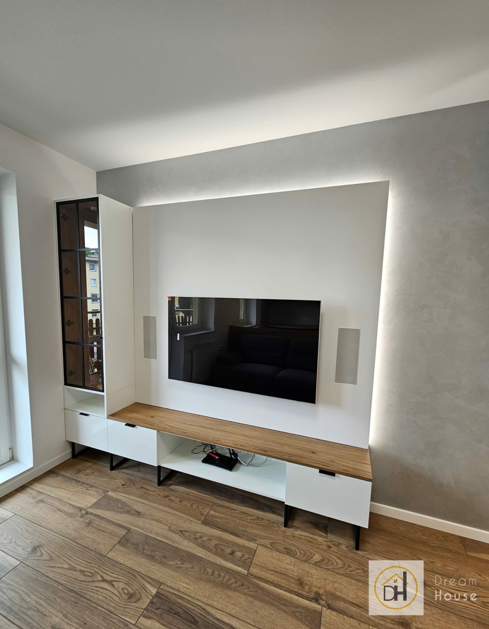 Nowoczesny salon z białymi meblami, drewnianym blatem i szarą ścianą z subtelnym oświetleniem LED. Telewizor wkomponowany w ścianę, drewniana podłoga.