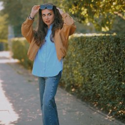 KarinaSafarova - Kobieta w brązowej kurtce i jeansach pozuje na chodniku, trzymając okulary na głowie. Za nią żywopłot i drzewa w słoneczny dzień.