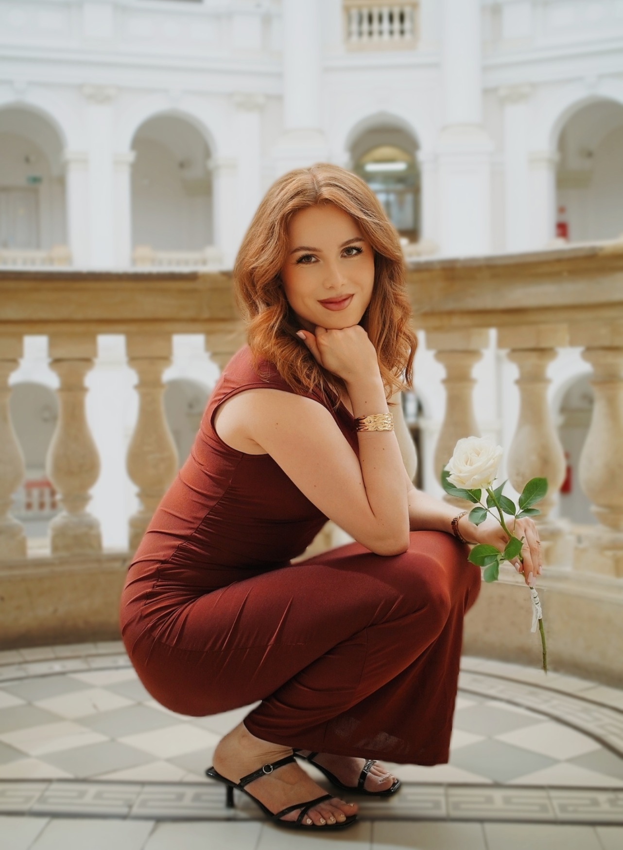 Kobieta w burgundowym kombinezonie kuca z białą różą na tle białej architektury z kolumnami i łukami. Uśmiech, bransoletka, wzorzysta podłoga.