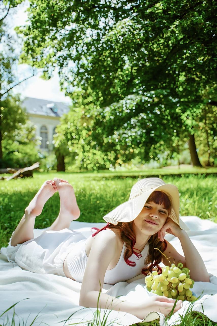 Młoda kobieta w kapeluszu leży na kocu w parku, trzymając kiść winogron. W tle zieleń drzew i budynek.